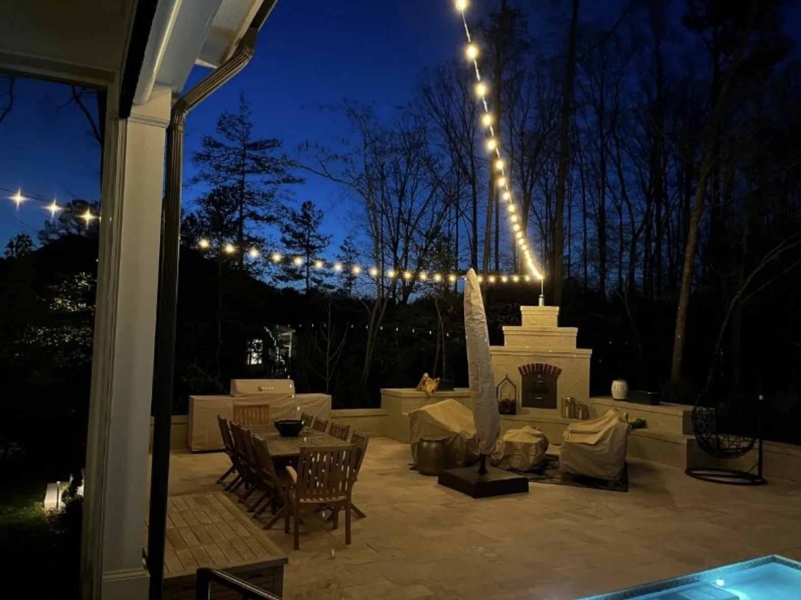 Bistro string lights over a backyard patio with outdoor fireplace and dining table